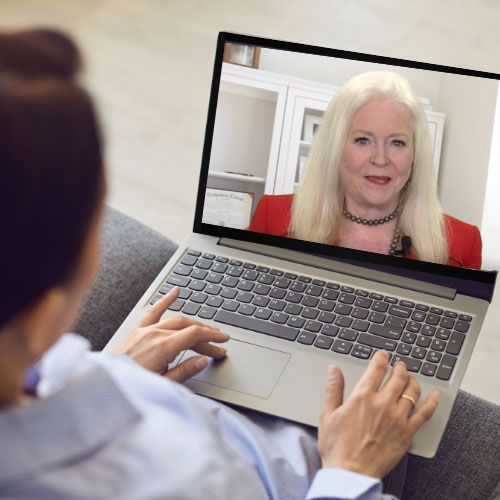 Person sitting with a laptop watching Sharon Lechter speak on screen, wearing a red blazer and black necklace in a well-lit office setting