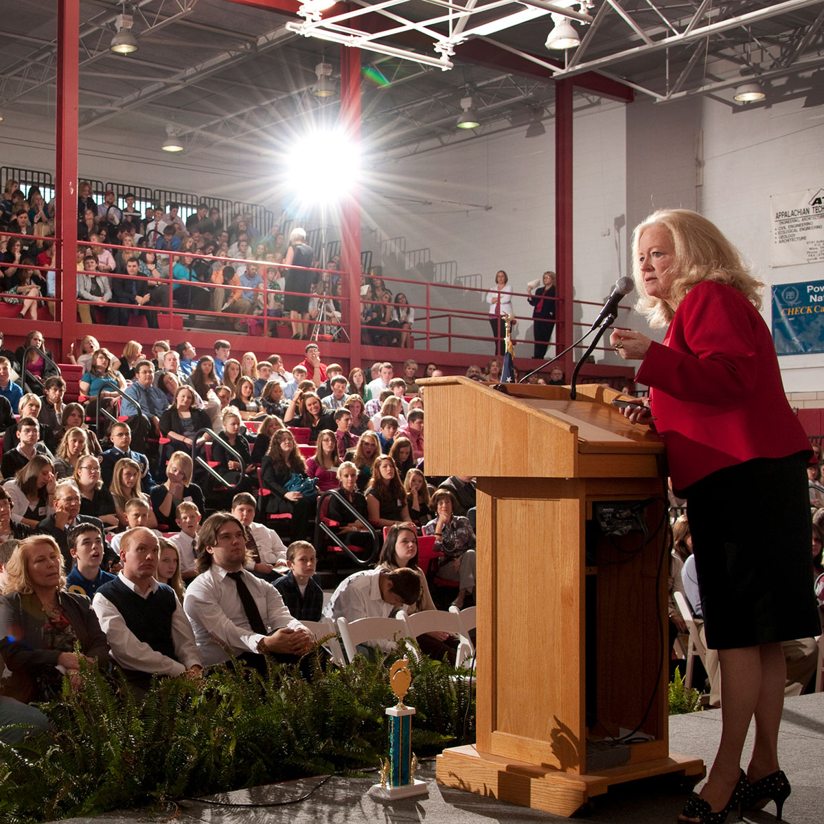 Sharon Lechter speaks at a podium in a crowded gymnasium. Behind her, a banner reads "Napoleon Hill Day.”