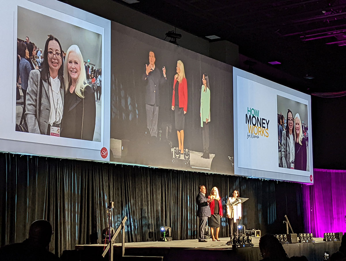 Sharon Lechter on stage at a conference with two other speakers, large screens behind them displaying event branding ‘How Money Works for Women’ and photos of Sharon with another attendee