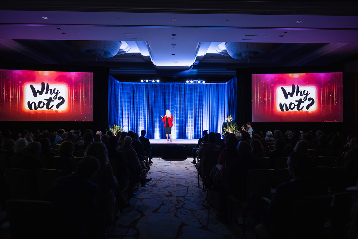 Sharon Lechter speaking on stage in front of a seated audience, wearing a red blazer and black dress, with large screens on either side displaying the phrase ‘Why not?’ against a colorful background.