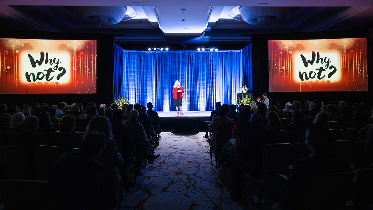 Sharon Lechter speaks on stage with a blue curtain backdrop in the middle of two screens displaying Why not