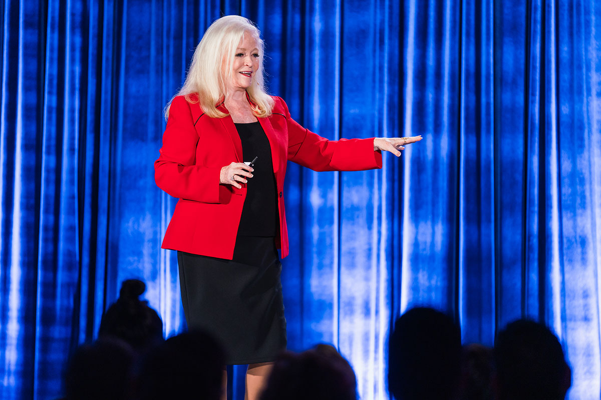 Sharon Lechter in a red blazer smiles while speaking on stage against a blue curtain