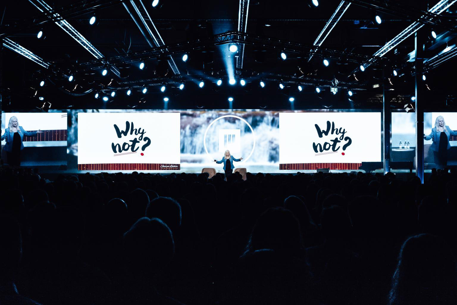 Sharon Lechter speaking on stage with a blue blazer in the middle of two screens displaying "Why not?