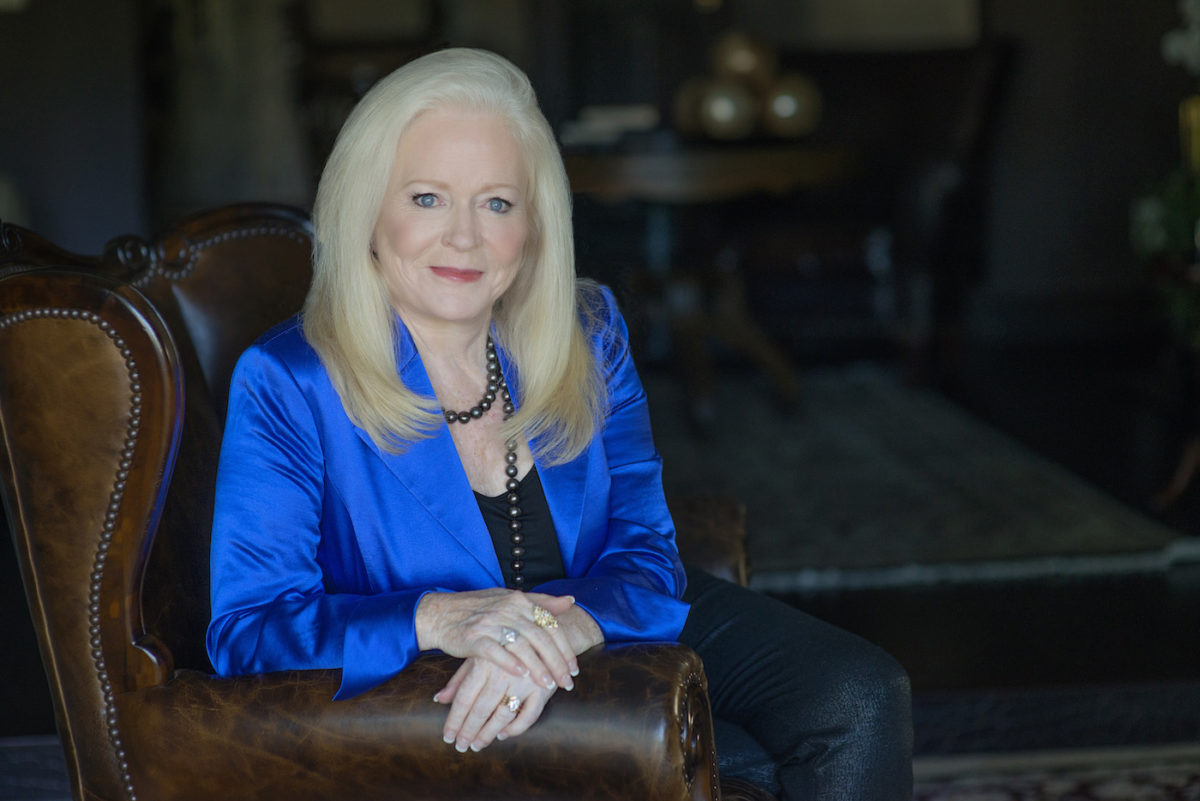 A confident woman with long blonde hair, in a blue blazer and black top, sits smiling on a brown leather chair. She wears a beaded necklace and rings, embodying the spirit of the Think and Grow Rich for Women Mastermind in a softly lit room.