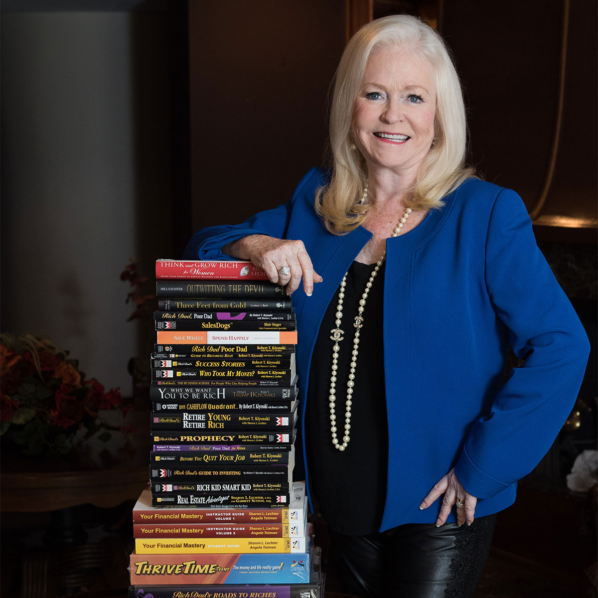 Sharon Lechter leaning on her stacked pile of books showcasing her own books and games