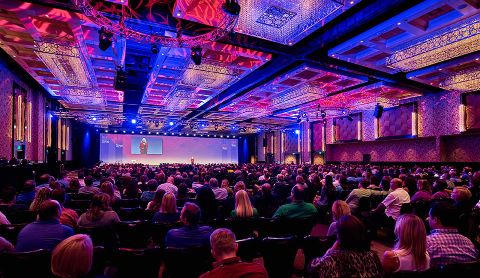 A large conference room filled with people watching Sharon Lechter speak on stage