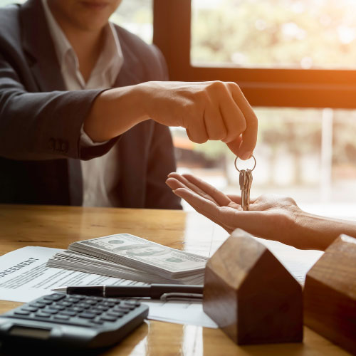 Person handing over house keys to another person across a desk with a contract, stack of cash, calculator, and small wooden house models