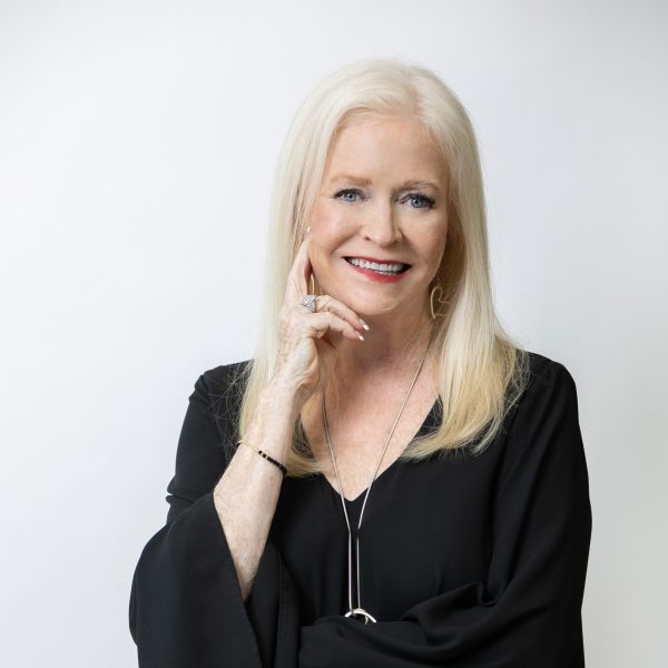 A smiling older woman with long blonde hair and silver jewelry stands against a white background, holding "Investing In Uncertain Times: Overcoming Fear and Embracing Opportunity," radiating confidence as she touches her face.