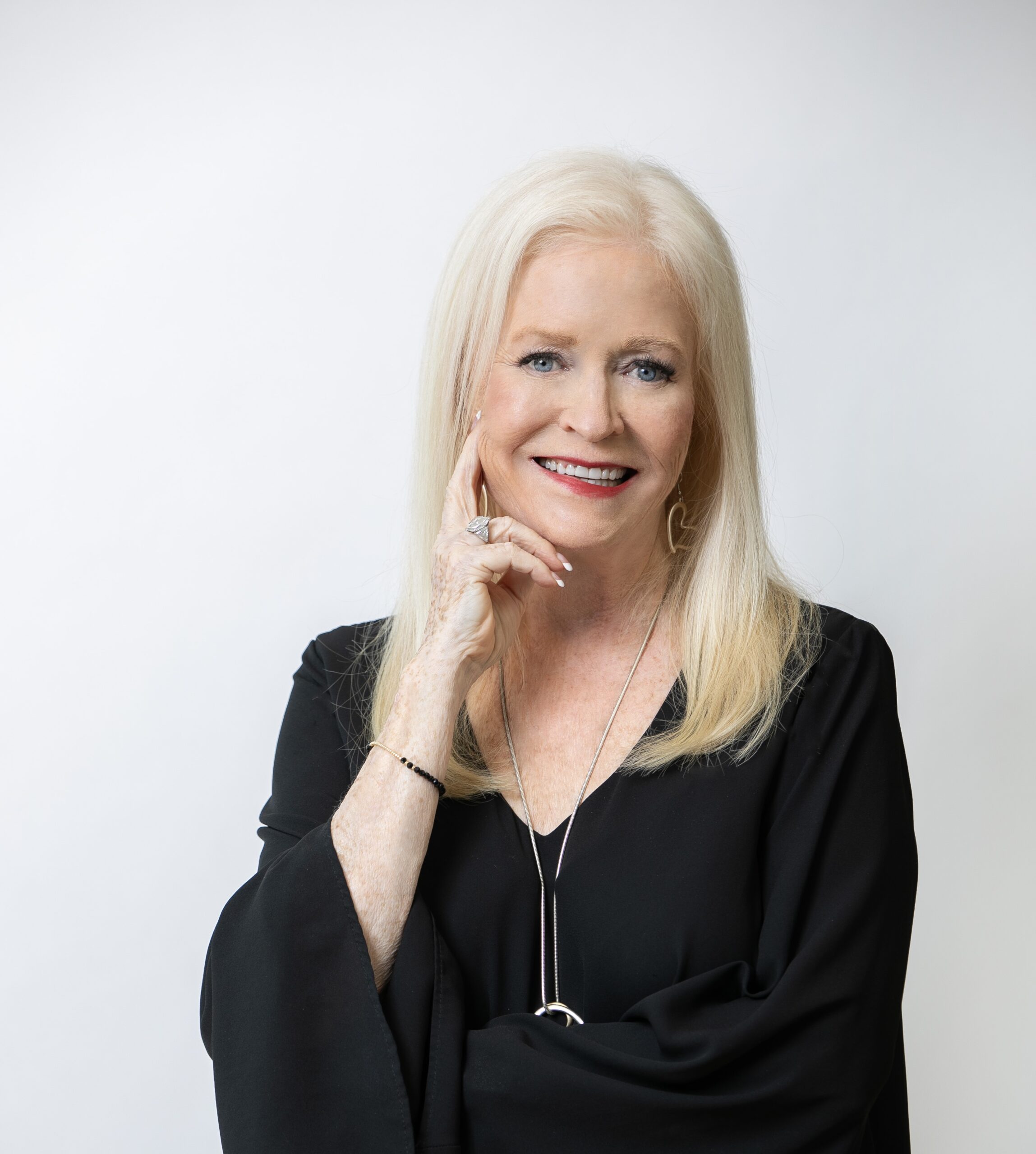 A smiling older woman with long blonde hair and silver jewelry stands against a white background, holding "Investing In Uncertain Times: Overcoming Fear and Embracing Opportunity," radiating confidence as she touches her face.