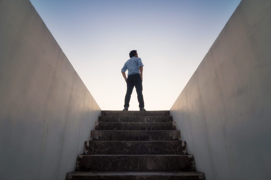 A person stands confidently at the top of a stone stairway, silhouetted against a clear sky