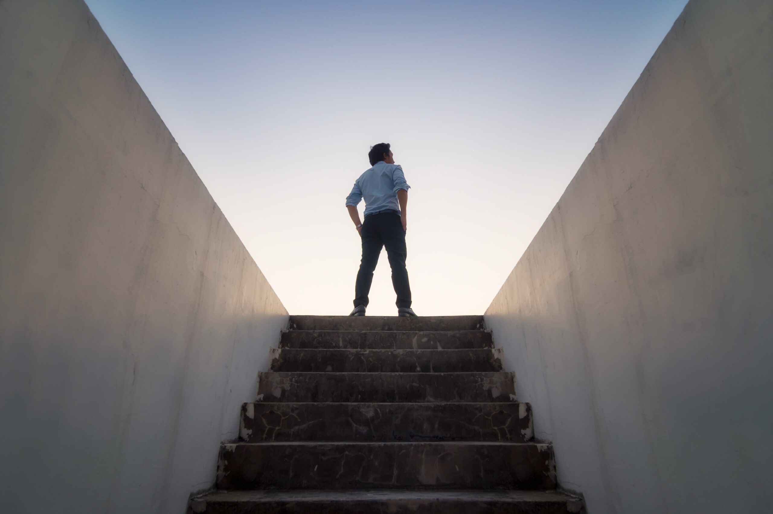 A person stands confidently at the top of a stone stairway, silhouetted against a clear sky