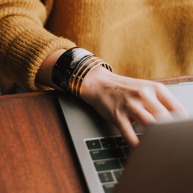 Someone types on a laptop while wearing a mustard yellow sweater and chunky bracelet at a wooden desk, taking the Play Big Financial Freedom and Entrepreneurial Course.