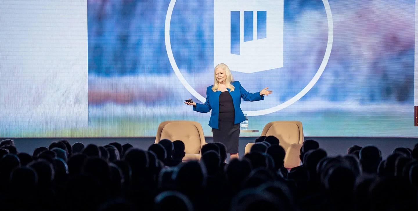 Sharon Lechter in a blue blazer speaking on stage, arms outstretched, in front of a large audience.