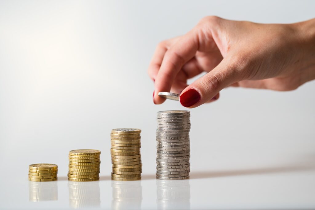 A hand with red nail polish adds a coin to one of four ascending stacks, symbolizing growth or investing.