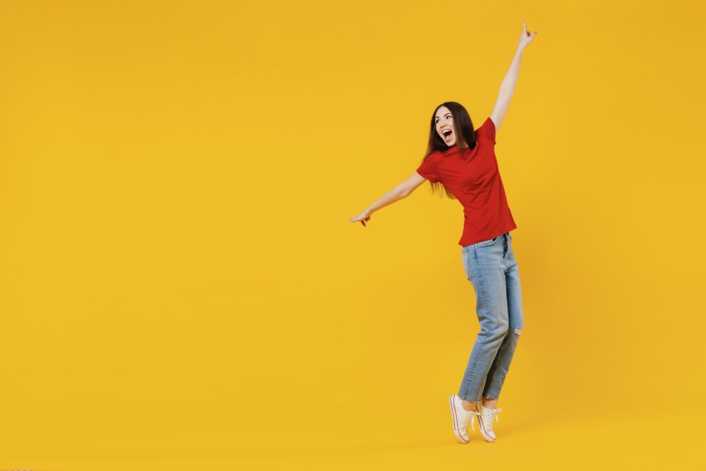 A woman in a red shirt and jeans joyfully jumps against a vibrant yellow background
