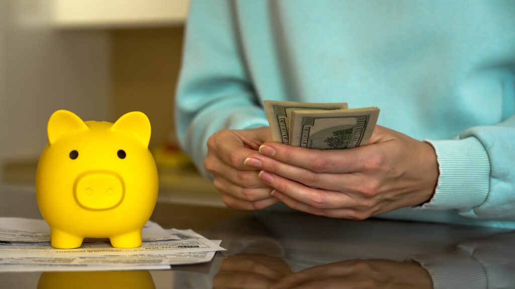 A person counting U.S. dollar bills next to a yellow piggy bank and some papers on a reflective surface, suggesting saving or managing money.
