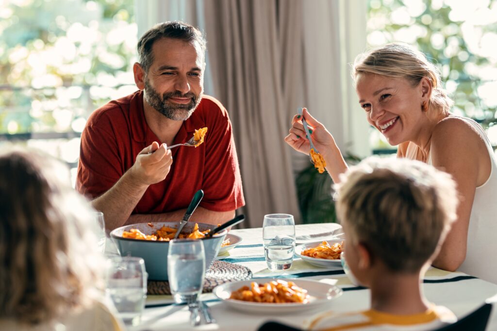 A smiling family of four sits around a dining table enjoying pasta, with sunlight streaming in. Two adults and two children are engaged in conversation, creating memories and building a long lasting legacy together.