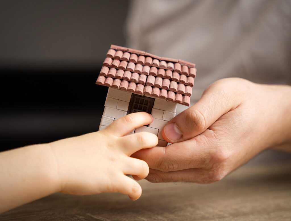 An adult hand and a child’s hand holding a small model house together