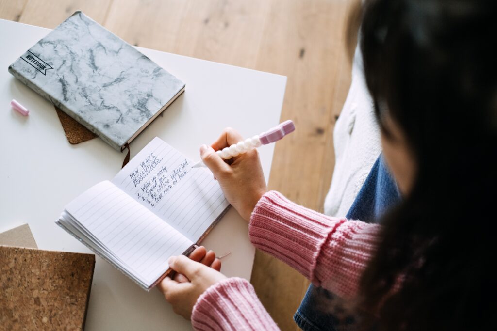 Person writing in a spiral notebook with a pastel pen on a white table.