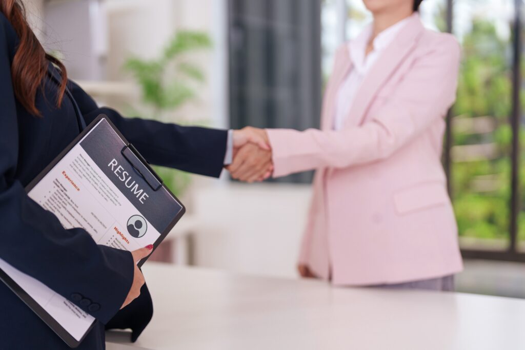 Two people in business attire are shaking hands across a table; one person is holding a resume folder