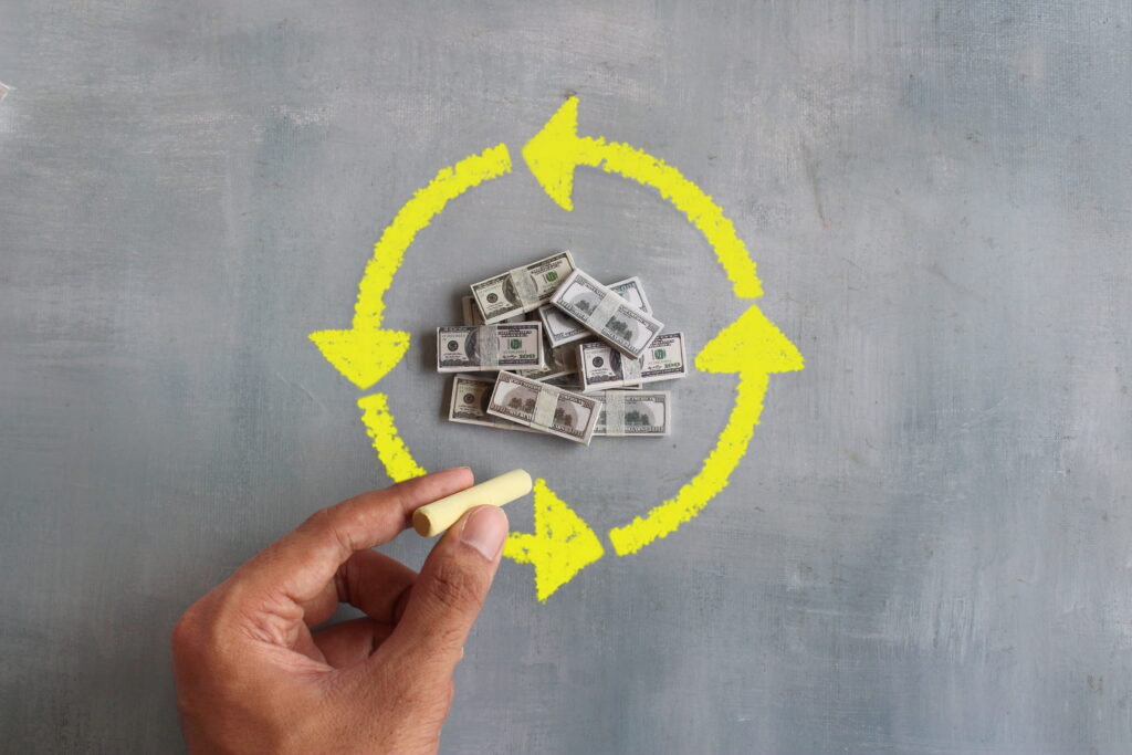 A hand holding yellow chalk draws a circular arrow around a pile of dollar bills on a gray background, symbolizing the concept of money circulation or financial flow.