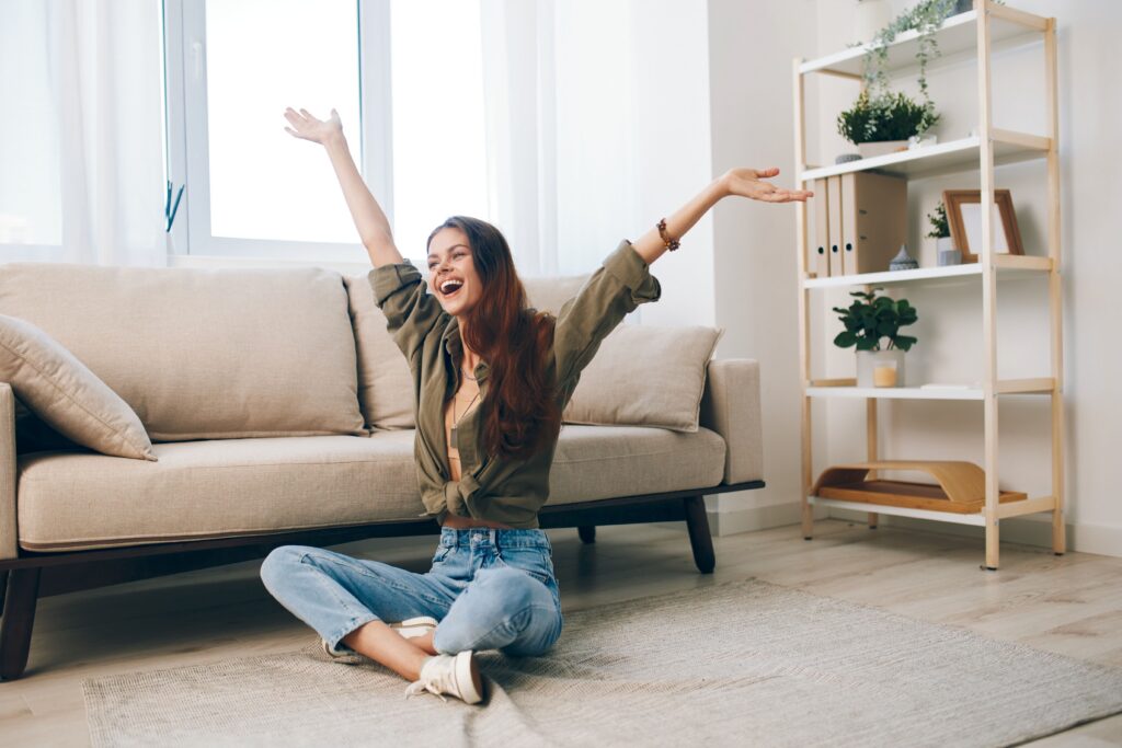 A young woman sits cross-legged on a rug in a bright living room, smiling with her arms raised in joy.
