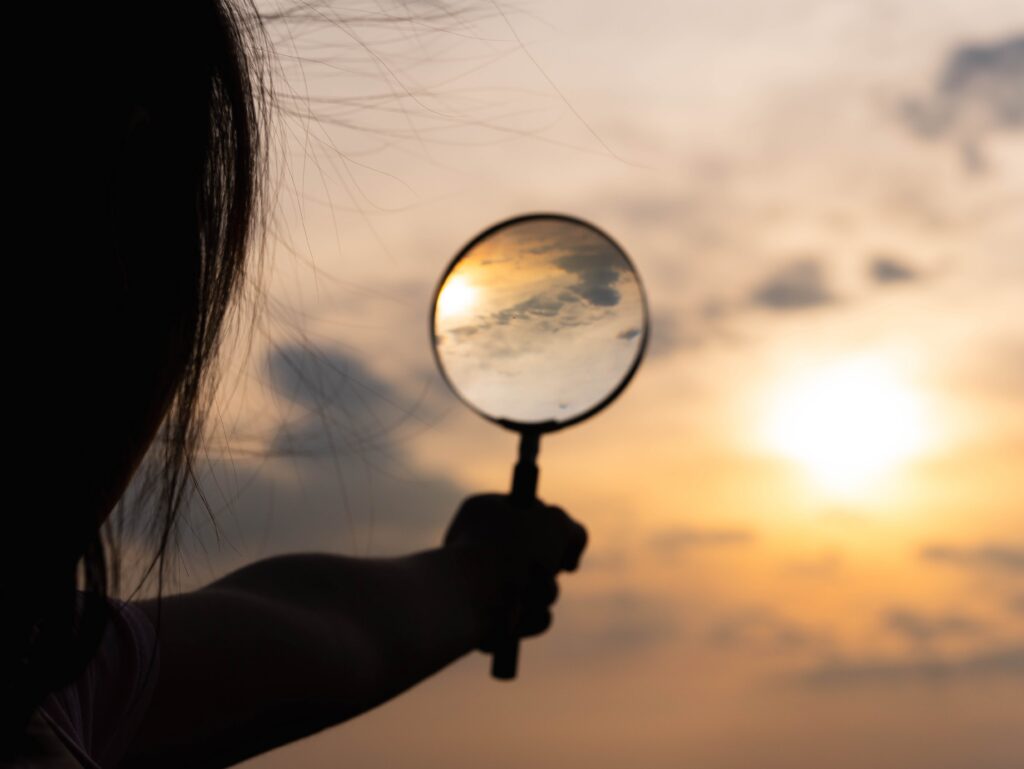 A person holds a magnifying glass toward the sky at sunset, with the sun and clouds visible through the lens.