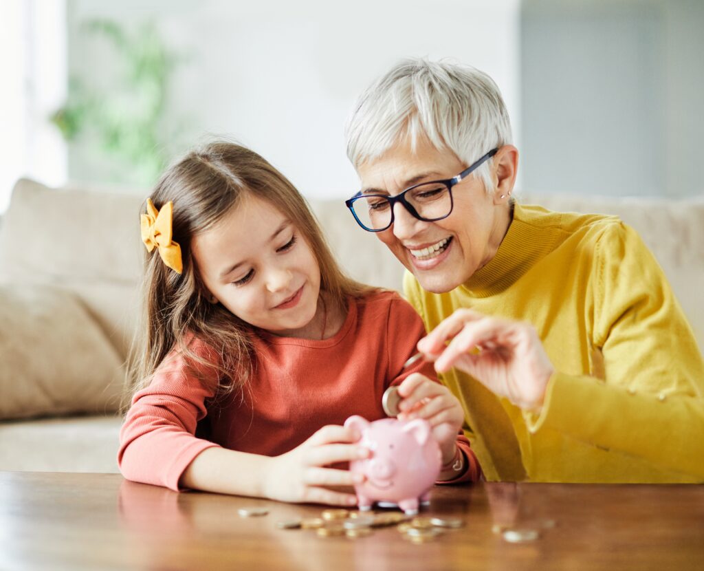 An older woman and a young girl smile as they put coins into a pink piggy bank on a table, teaching about saving money.