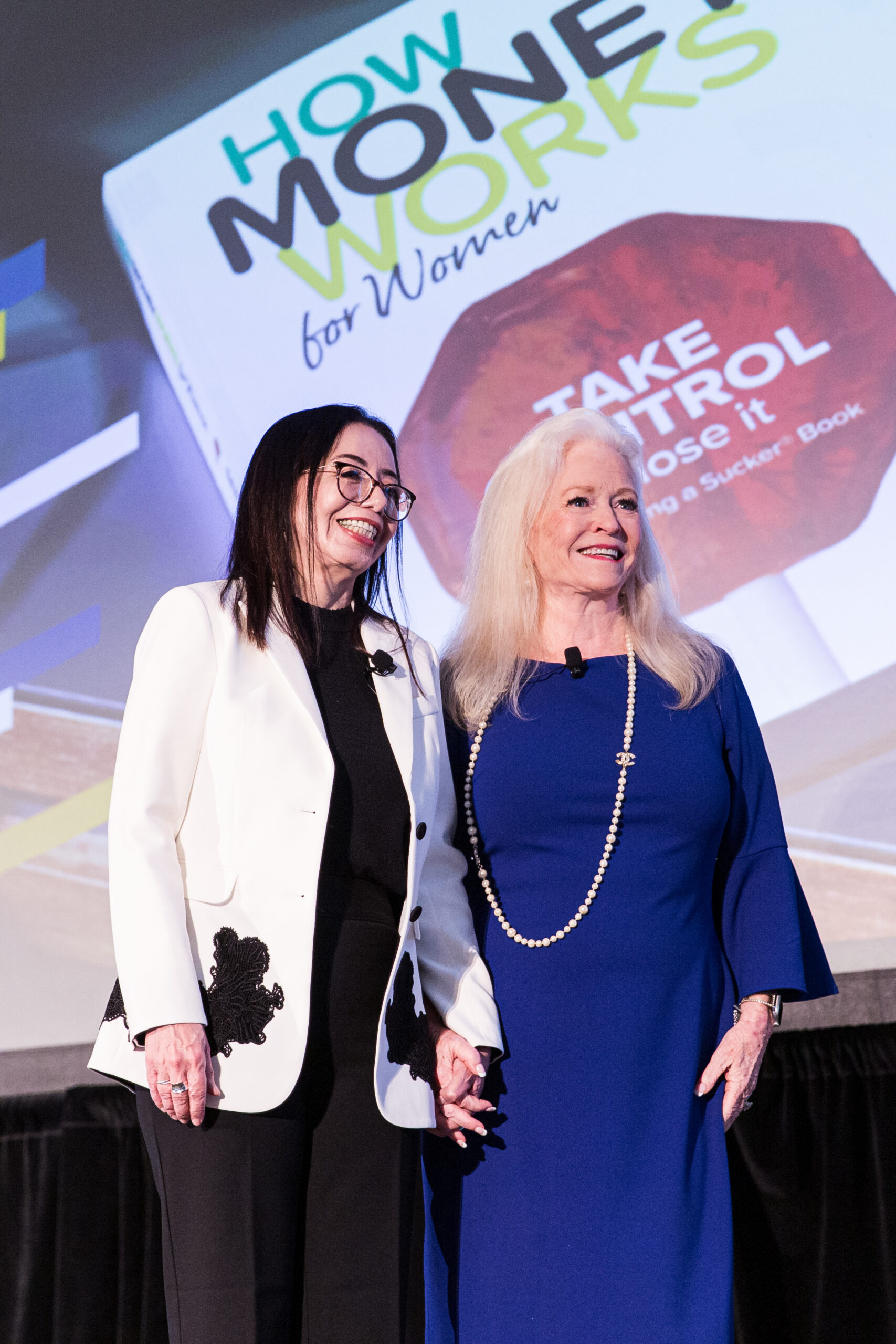 Two women stand smiling on stage, holding hands. Behind them is a large screen featuring the book cover How Money Works for Women with the phrase Take Control visible.