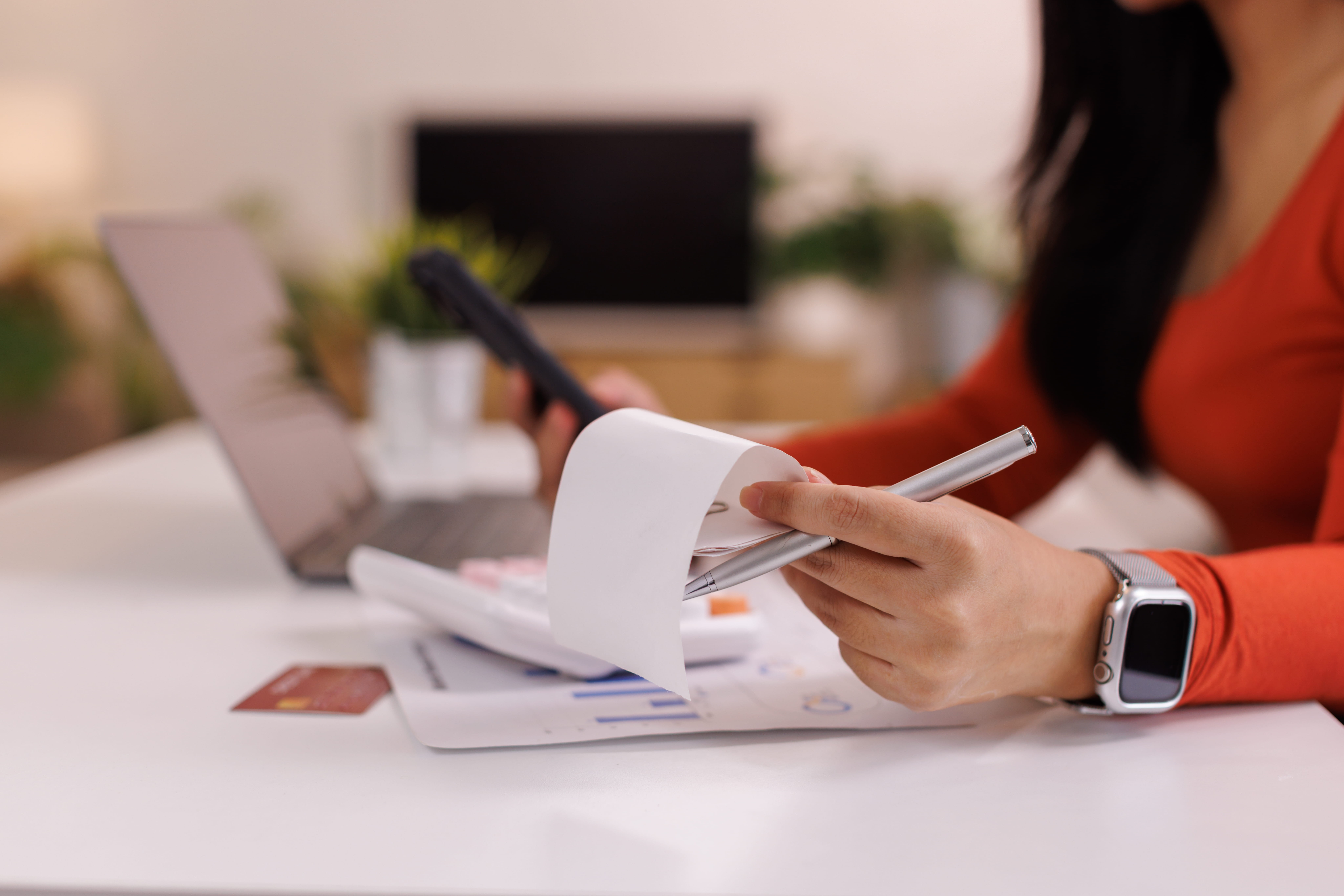 A woman wearing a smartwatch reviews receipts and paperwork while holding a pen at a desk with a laptop, documents, and a credit card. The background is softly blurred.